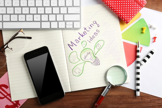 Um caderno sobre um fundo de madeira apresenta o texto 'Ideias de Marketing' ao lado de um desenho de uma lâmpada.