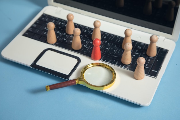 Uma lupa está posicionada sobre figuras humanas de madeira colocadas em um teclado de laptop, simbolizando o processo de seleção de candidatos.