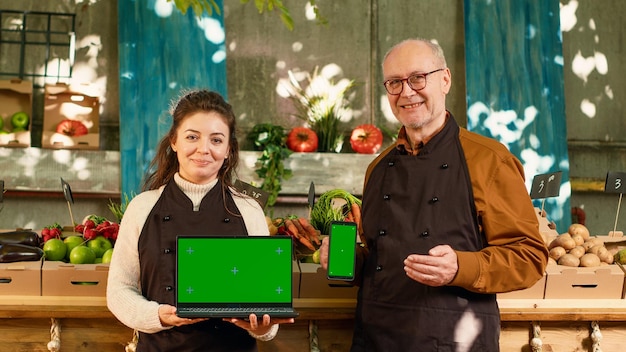Um homem e uma mulher, proprietários de negócios em uma feira de agricultores, seguram uma tela verde em um laptop e em um celular, apresentando um espaço em branco para conteúdo digital.
