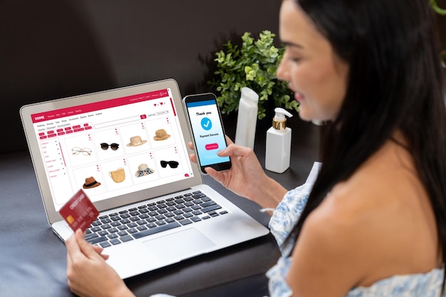 Uma jovem mulher está sentada em uma mesa usando um notebook para fazer compras online, enquanto segura um smartphone para realizar o pagamento com um cartão de crédito.